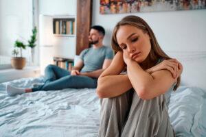 Junge Frau, die enttäuscht und traurig auf dem Bett sitzt, im Hintergrund sitzender Mann, Symbol für familiäre Probleme.